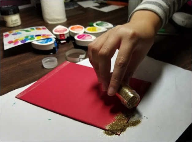 A child applies glitter to a taped area of cardstock in the process of making her handmade Christmas card.